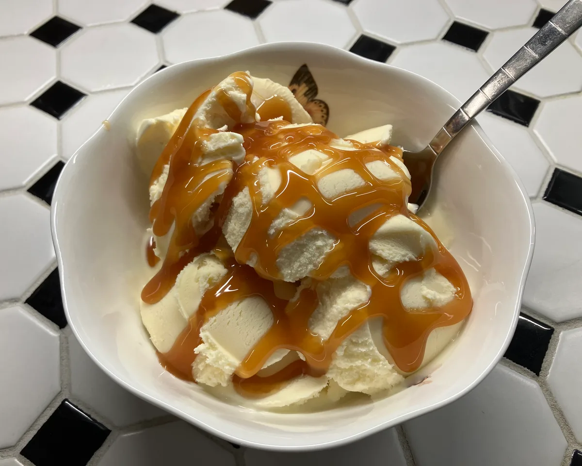 Bowl of vanilla ice cream drizzled with caramel syrup on a black and white tile counter, with a spoon resting in the bowl.