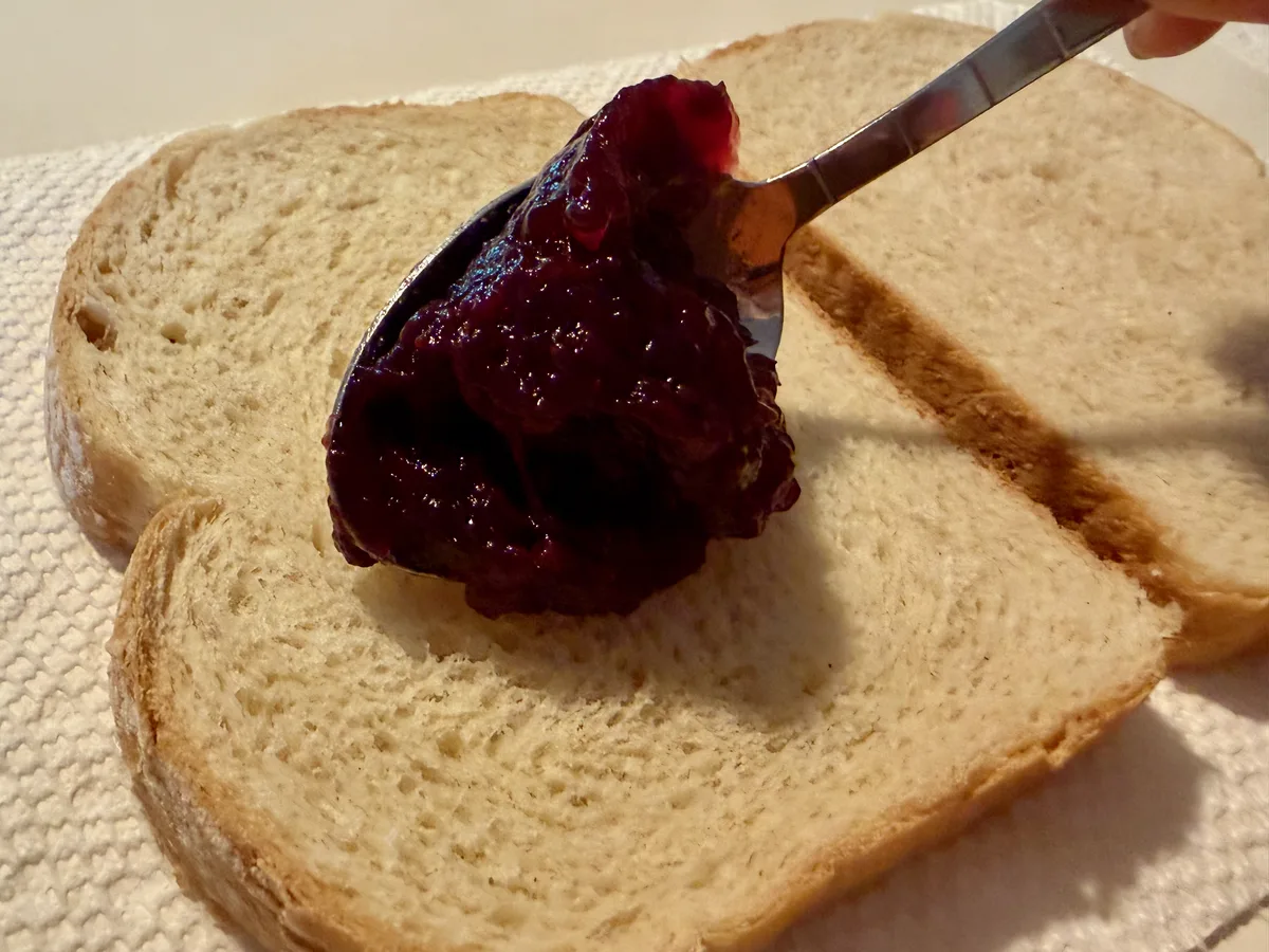 A spoonful of berry fruit spread being placed onto a slice of sourdough bread.