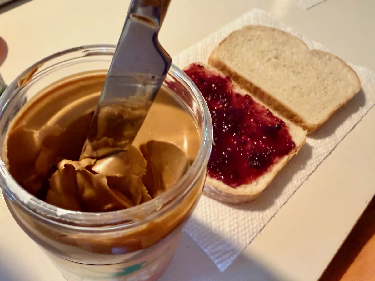 A knife dipped into a peanut butter jar, beside a jam-covered slice of bread.