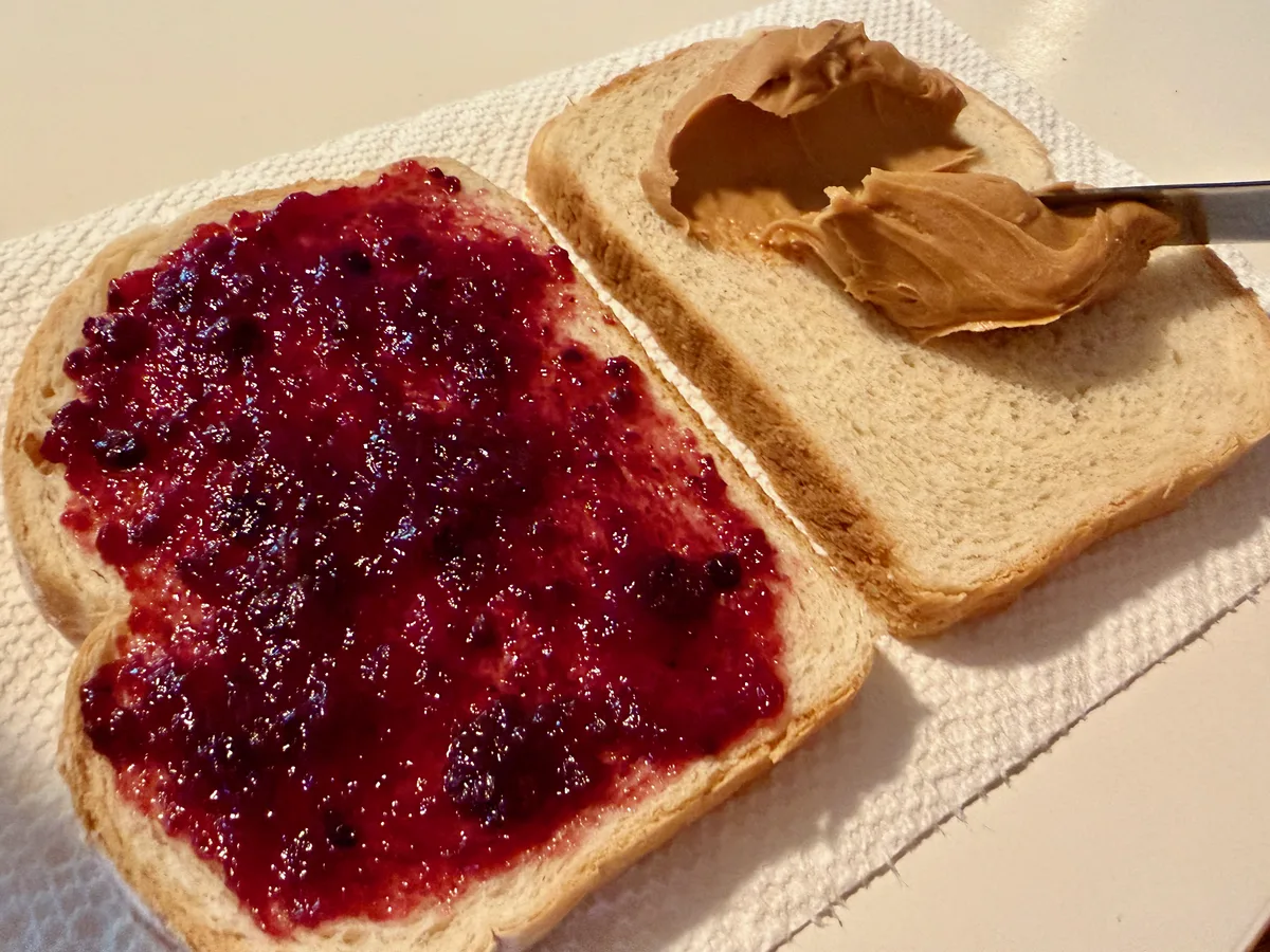 A slice of bread with peanut butter being spread, next to a finished jam slice.