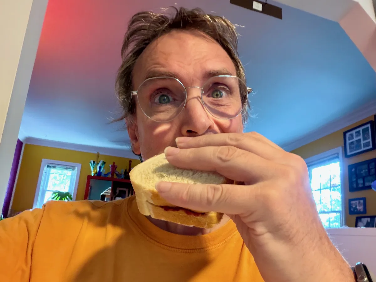 Thad wearing glasses and an orange shirt about to bite into a sandwich.