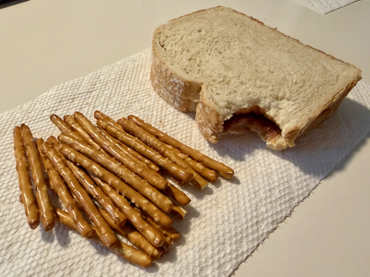 A bitten sandwich and a small pile of pretzel sticks on a paper towel.