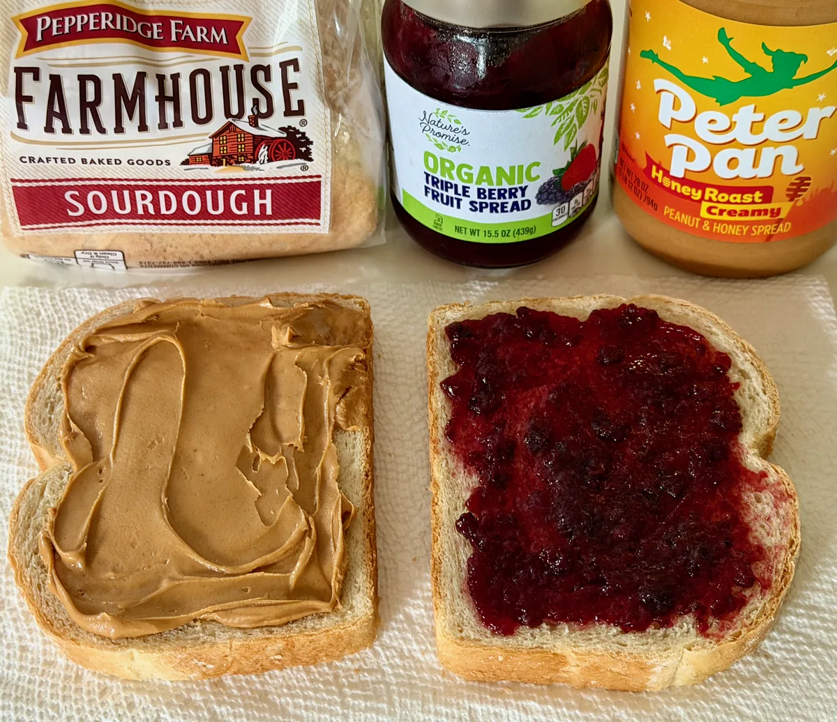 Two slices of bread with peanut butter and jam, next to sourdough bread, fruit spread, and peanut butter jars.
