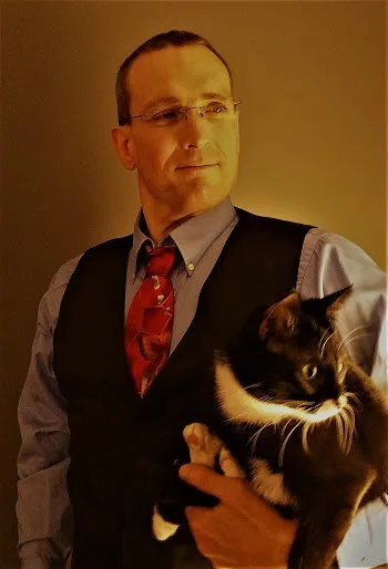 Thad in a vest and tie holding his tuxedo cat, Miss Muffin, in warm indoor lighting.