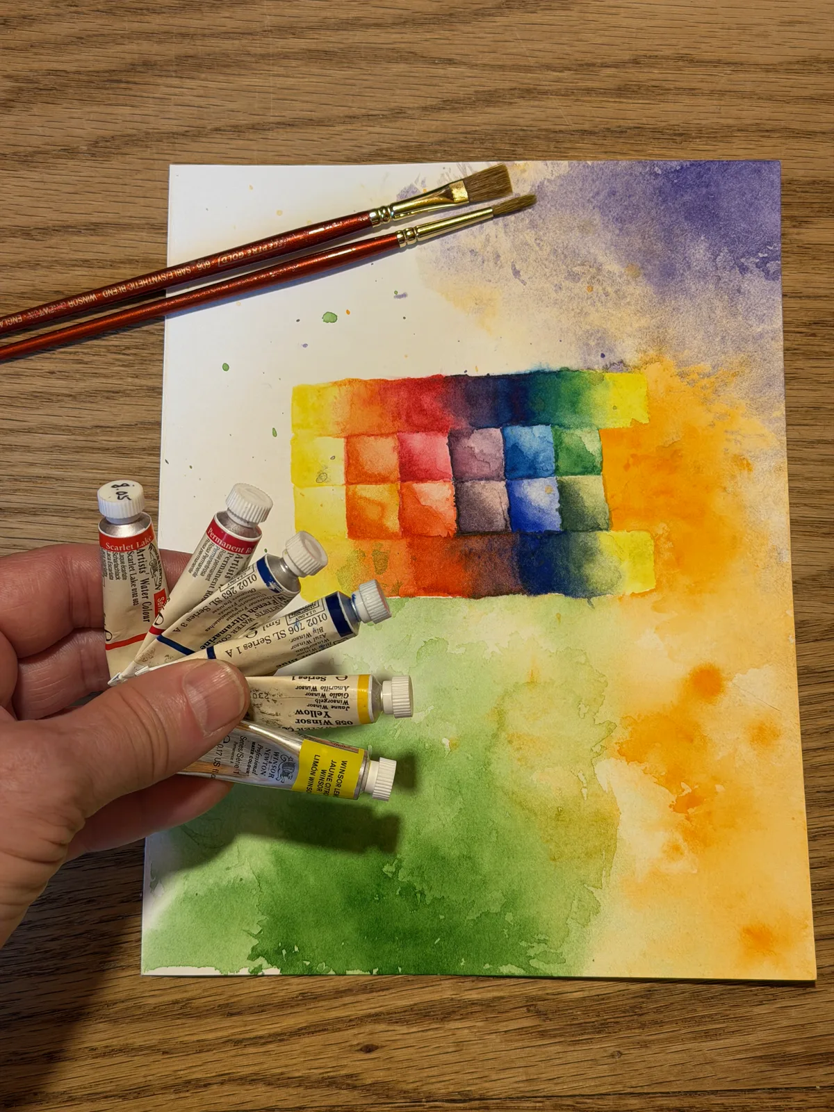 Watercolor tubes held over a finished palette study painting, with two brushes resting on the paper.