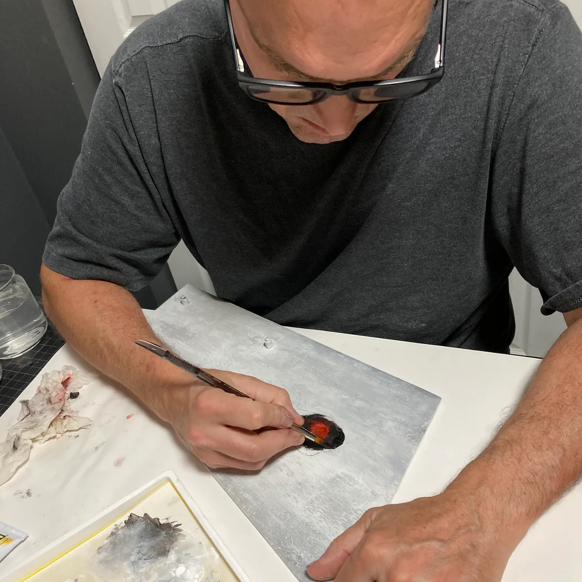 Thad using a brush to refine red paint applied first with his fingers on the gray underpainted wood panel.