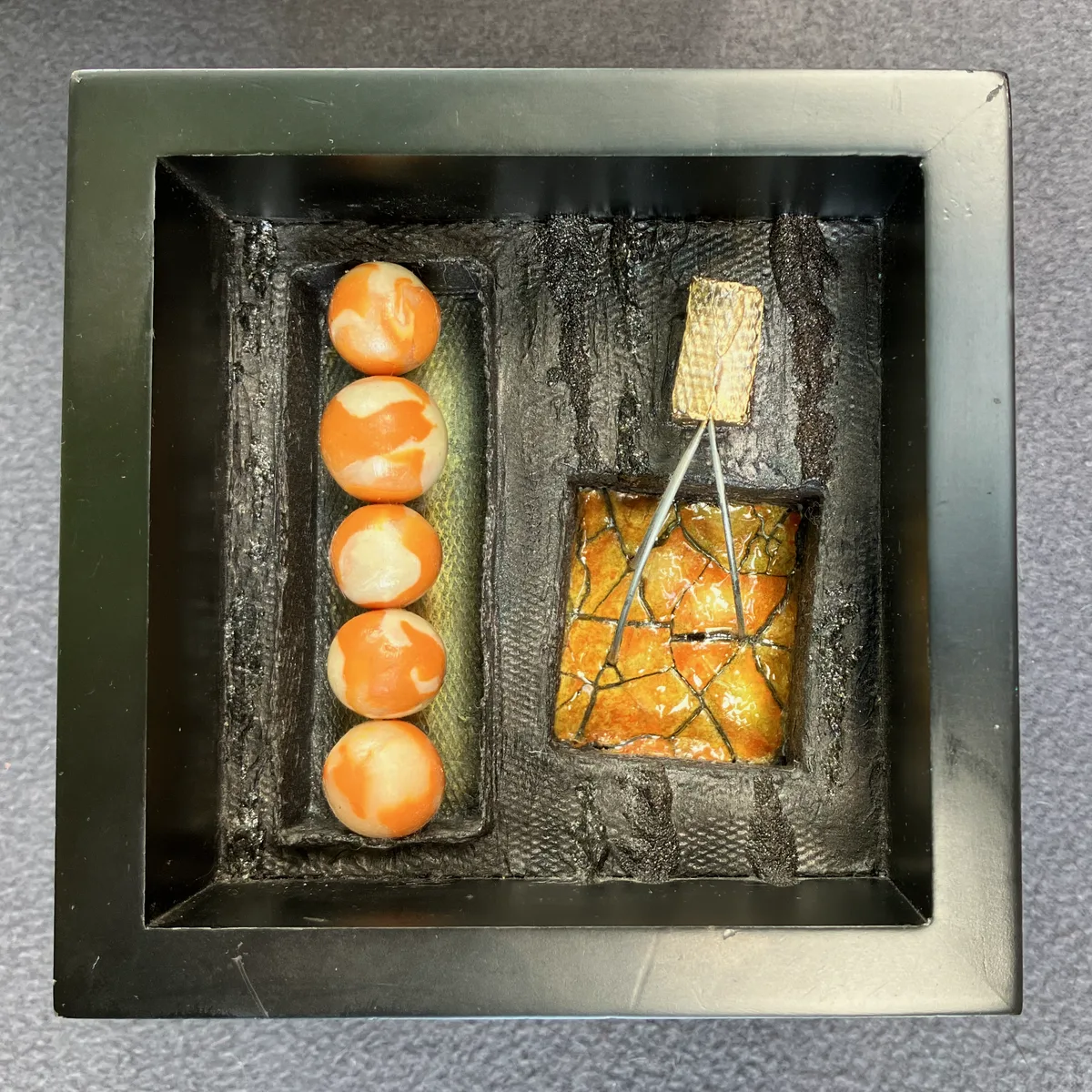 A small mixed media artwork combining five marbled clay beads and a textured gold square, mounted in a black shadow box.