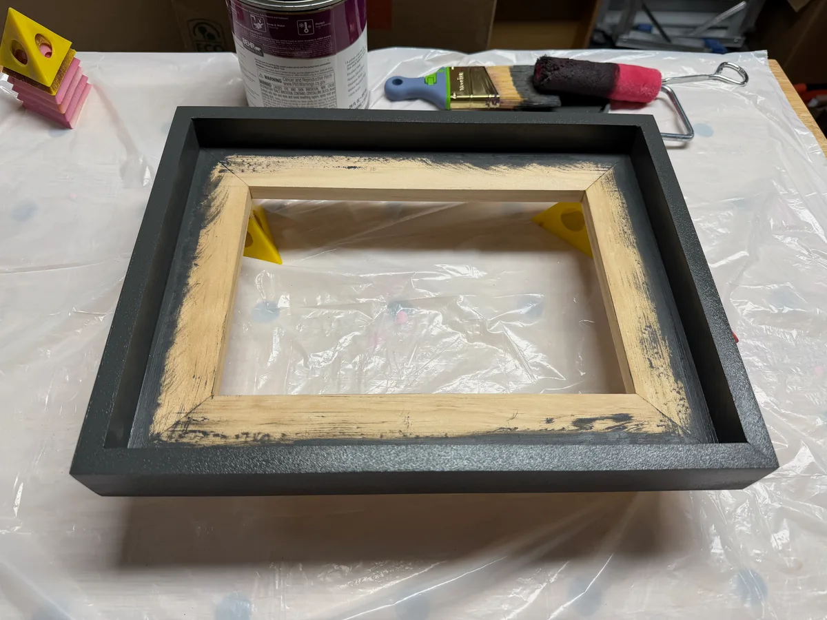 The wood frame partway through painting, black on the outer edge, raw wood still visible along the inner lip.