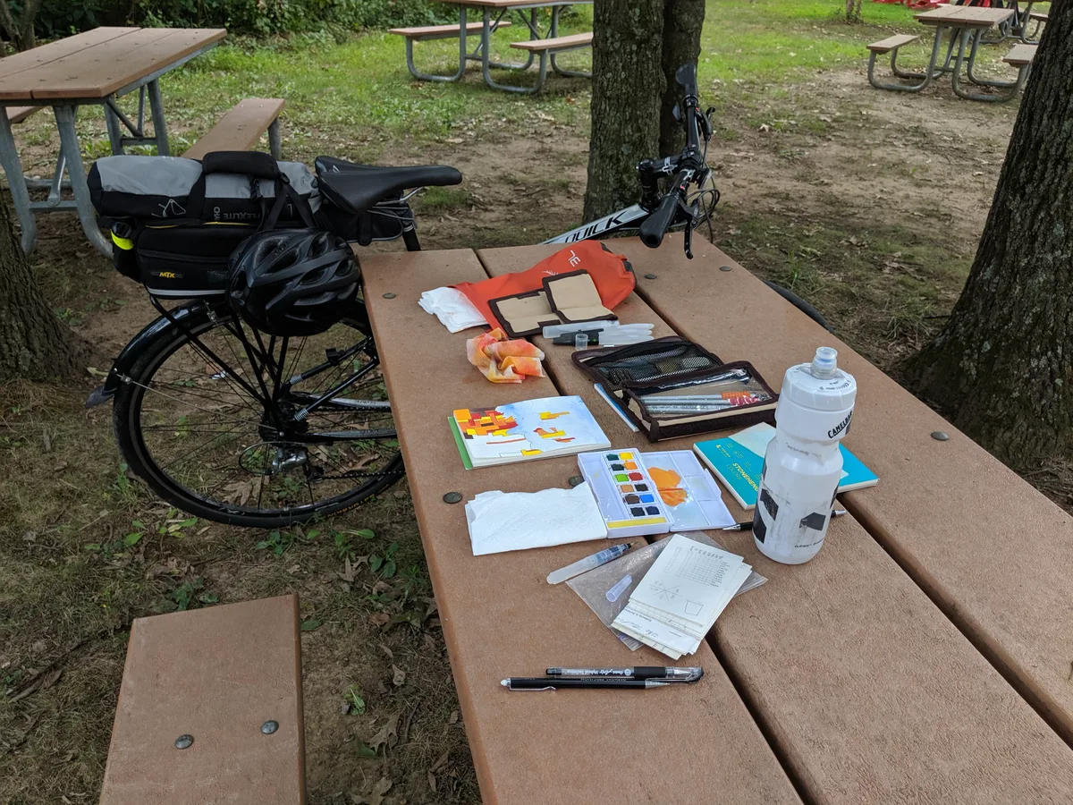 A travel watercolor kit and sketchbook spread across a picnic table, bicycle leaning against the bench.